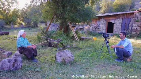 Der Journalist Saba Tsitsikashvili im Interview mit einer Dorfbewohnerin an der Grenze zu Südossetien: Sie sitzen sich mit etwas Abstand draußen auf Baumstümpfen vor einer gemauerten Hütte und einem Baum gegenüber. Der Journalist filmt mit einer Videokamera.